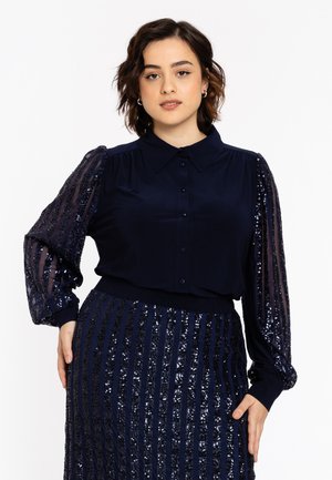Kvinde med kort mørkt hår iført navybluse med gennemskinnelige pufærmer med palietter og matchende palietskørt, stående med den ene hånd på hoften.