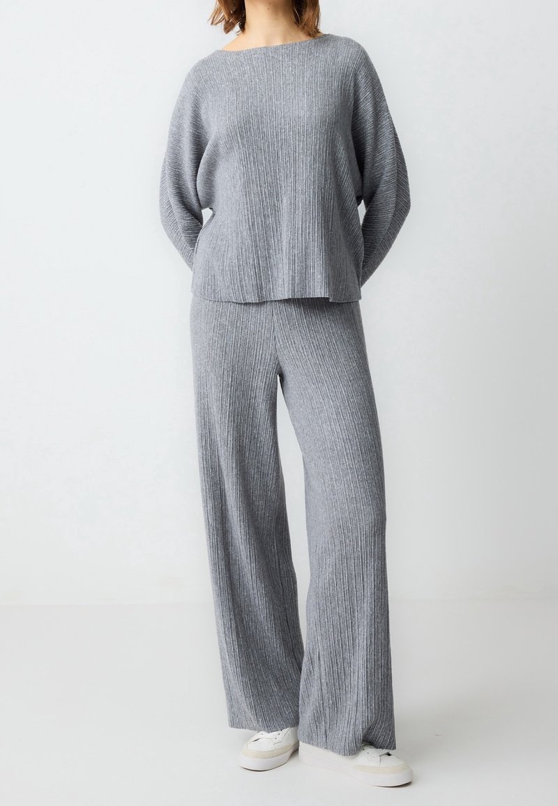 Femme portant un pull en maille côtelée gris ample et un pantalon large assorti avec des baskets blanches, debout devant un fond clair uni.