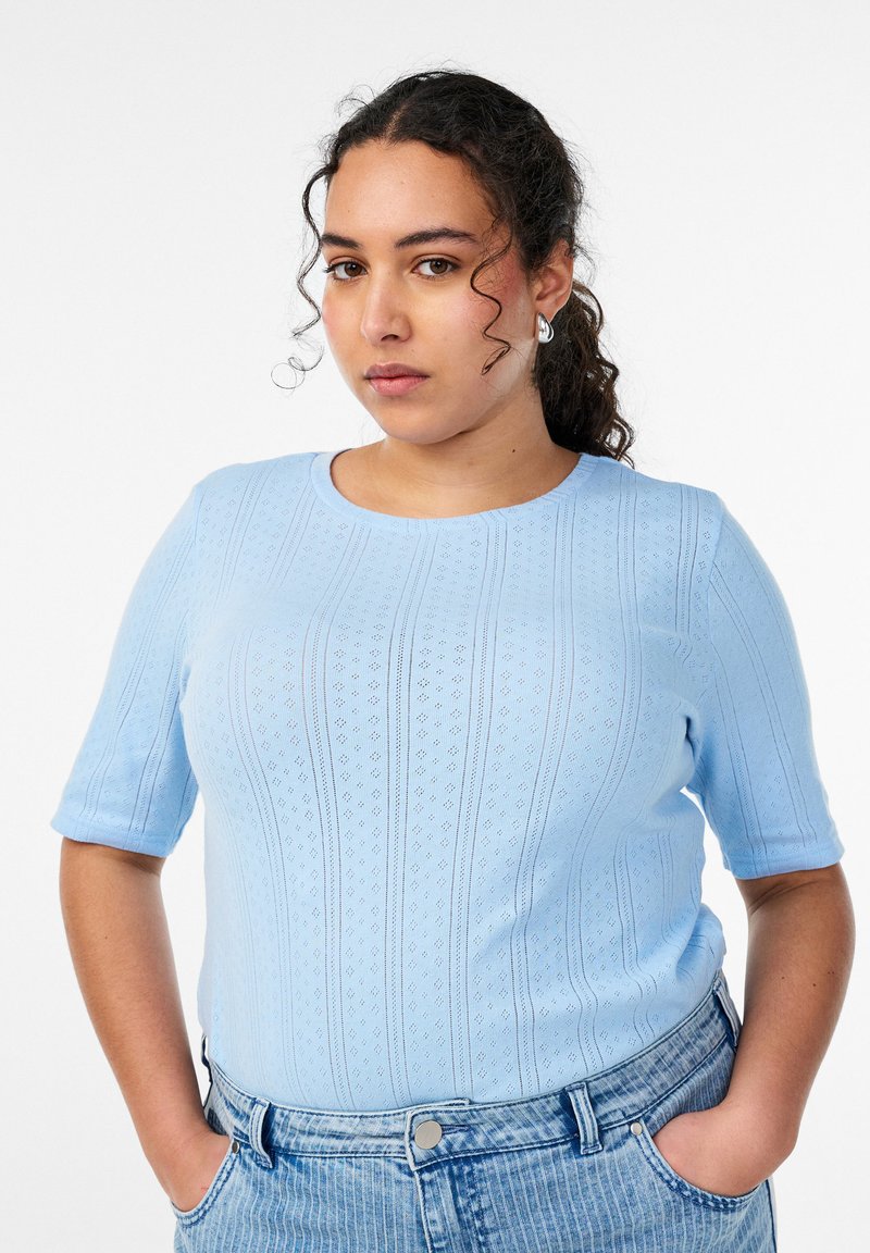 Mlada žena s kovrčavom kosom, odjevena u svijetlopludu pletenu majicu i traperice visokog struka, stoji s rukama u džepovima na bijeloj pozadini.