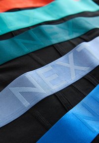 Quatre paires de sous-vêtements noirs pliés avec de larges ceintures de taille colorées en orange, turquoise, bleu clair et bleu, chacune embossée du mot « NEXT ».