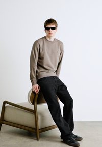 Sudadera marrón con cuello redondo, pantalones negros y gafas de sol oscuras, sentado en una silla de madera con asiento acolchado. Fondo claro.