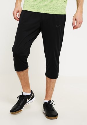 Mann trägt schwarze kniehohe Nike-Sporthose, schwarz-weiße Nike-Sneaker und ein hellgrünes Shirt, steht auf hellem Boden.