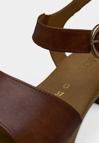 Καφέ σανδάλι από συνθετικό δέρμα με φαρδιά λουράκι, στρογγυλό τρουκ και μαλακό πάτο. Διαθέτει χαμηλό τακούνι σε στιλ στοίβας.