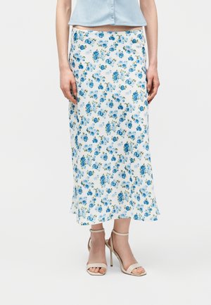 Kobieta ubrana w białą spódnicę midi z niebieskim kwiatowym wzorem, jasnoniebieską koszulę zapinaną na guziki oraz beżowe sandały na wysokim obcasie, stojąca w pomieszczeniu.