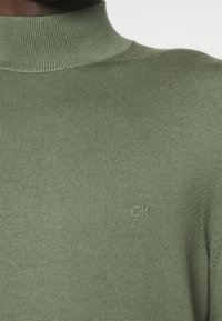 Maglione a collo alto verde a costine realizzato in tessuto testurizzato, con logo CK ricamato sul petto. Design semplice e aderente.