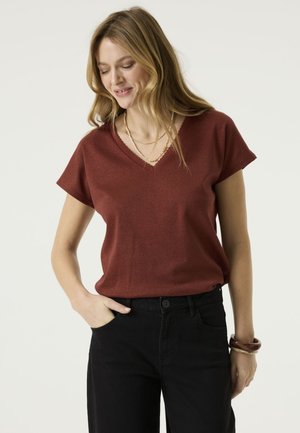 Vrouw met lang blond haar die een roestkleurige korte mouw V-hals top draagt, zwarte broek, meerdere kettingen en armbanden op een lichte achtergrond.