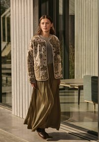 Giacca beige ricamata con colletto rotondo, abbinata a una gonna fluida verde oliva. Il modello indossa stivali marrone scuro. Ambientazione interna minimalista.
