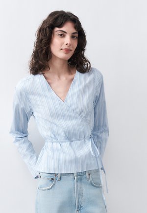 Νεαρή γυναίκα με σγουρά μαλλιά, που φοράει ένα ανοιχτό μπλε ριγέ μπουφάν τύπου wrap και ανοιχτόχρωμα τζιν, στέκεται μπροστά σε ένα απλό φόντο.