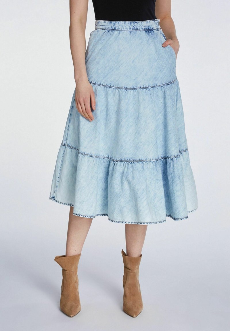 Jupe en denim bleu clair à volants avec un ourlet effiloché, dotée de poches et de surpiqûres contrastantes. Associée à des bottines beige à la cheville.