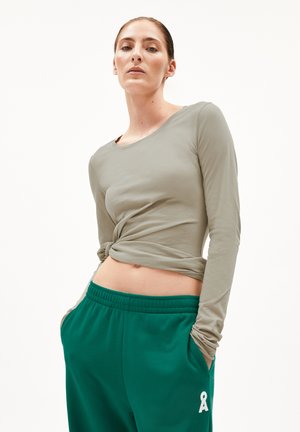 Langärmliges, hellolivfarbenes Hemd mit geknotetem Frontdetail, kombiniert mit grünen Sweatpants, die auf der linken Seite ein weißes Logo zeigen.