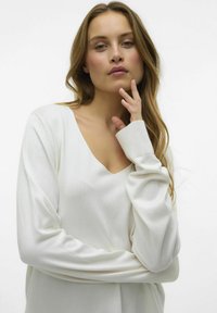Pull blanc à manches longues avec un design en V, présentant des poignets côtelés et une texture douce, légèrement oversize.