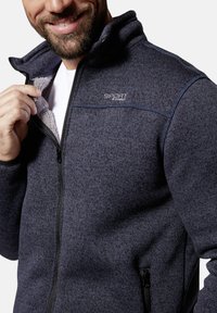 Marineblaue Fleecejacke mit hohem Kragen, durchgehendem Reißverschluss und Seitentaschen. Sie verfügt über einen strukturierten Stoff und ein dezentes Logodesign auf der Brust.
