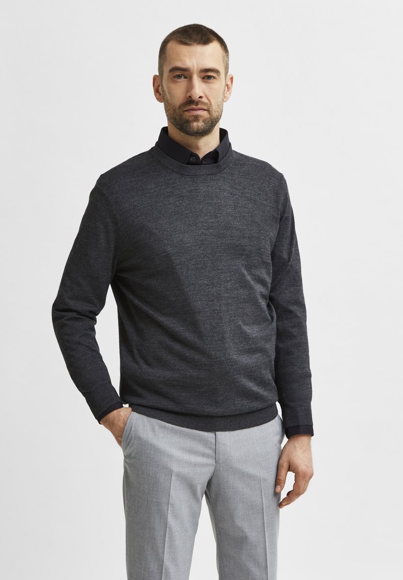 Dark grey knit sweater with a round neckline, worn over a black collared shirt. Paired with light grey tailored trousers, smooth texture.