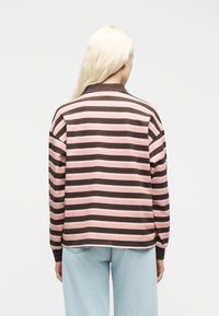 Άτομο με μακριά ξανθά μαλλιά που φοράει καφέ και ροζ ριγέ πουκάμισο με μακριά μανίκια και ανοιχτό μπλε τζιν, γυρισμένο πλάτη σε λευκό φόντο.