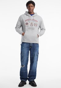 Felpa grigia con cappuccio e tasca frontale, impreziosita da loghi stampati, abbinata a jeans cargo blu scuri con effetto usurato e scarpe nere resistenti.