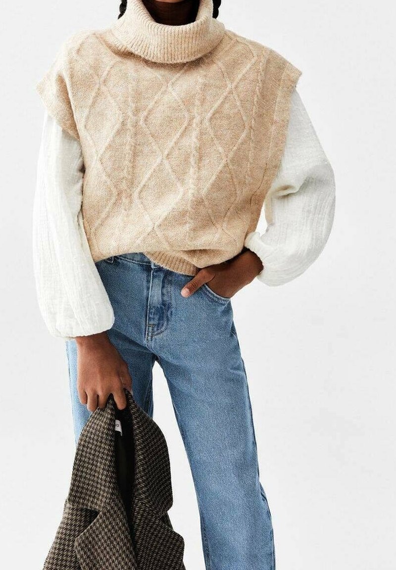 Personne portant un col roulé sans manches en maille torsadée beige sur un haut blanc à manches bouffantes, jean bleu, tenant une veste marron à motifs dans la main gauche.