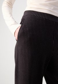 Personne portant un pantalon noir à fines rayures avec la main dans la poche et un haut à manches longues beige sur un fond blanc uni.