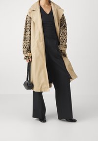 Beige vaelluskotelo mustilla kuviollisilla hihoilla, päällä musta V-pääntie pusero ja leveälahkeiset mustat housut. Mukana pieni musta olkalaukku.