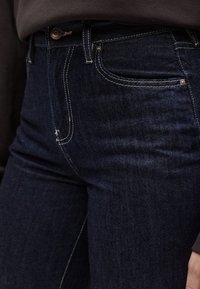 Sötétkék farmernadrág kontrasztos fehér varrással, egyenes szabású, kis elülső zsebbel és fém gombos záródással.