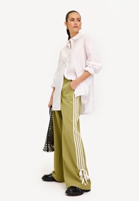 Witte oversized blouse, olijfgroene wijde broek met witte zijstrepen, gecombineerd met zwarte chunky schoenen en een zwarte gestructureerde tas.