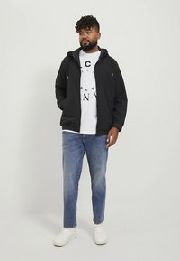 Black zip-up jacket with a hood over a white graphic tee, paired with light blue jeans and white sneakers on a plain background.