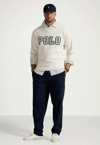 Άνδρας με ναυτικό καπέλο, κρεμ φούτερ με το κείμενο "POLO", ανοιχτό μπλε πουκάμισο, ναυτικού χρώματος παντελόνι, λευκά παπούτσια, στέκεται με τα χέρια στις τσέπες.