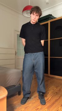 Zwarte kortademige polo, losse blauwe jeans en zwarte sneakers. Houten vloer en een rode hanglamp in een kamer met planken.