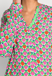 Blusa colorata con un motivo geometrico in rosa, verde, rosso e blu. Realizzata in morbido tessuto, con apertura a bottoni e scollatura a V.
