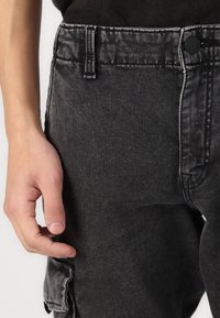Zwarte denim cargobroek met een knoopsluiting, riemlussen en contrasterend witte stiksels. Een hand rust nabij een zijzak.