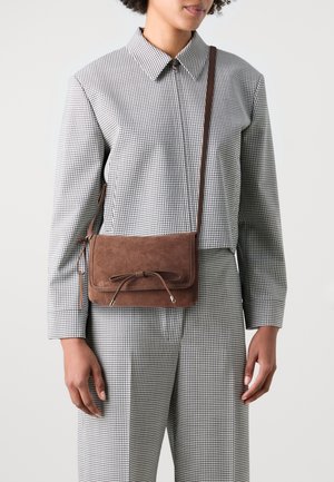 Femme portant un costume à carreaux noir et blanc, avec un sac bandoulière en daim marron orné d'un nœud sur le rabat avant.