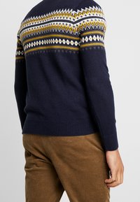 Marinblå stickad tröja med flerfärgad fair isle-mönstring på oket; ribbade muddar och nederkant; ihop med ljusbruna kordbyxor.