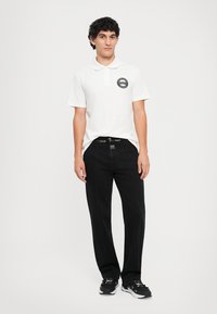 Λευκό πόλο μπλουζάκι με στρογγυλό μαύρο λογότυπο στο στήθος, συνδυασμένο με μαύρα τζιν παντελόνια κανονικής γραμμής με λευκές ραφές. Μαύρα αθλητικά παπούτσια ολοκληρώνουν το σύνολο.