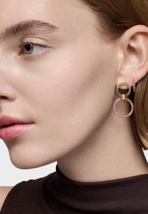 Gros plan du côté gauche du visage d'une femme et de son oreille portant des boucles d'oreilles doubles en or ornées de pierres précieuses multicolores.