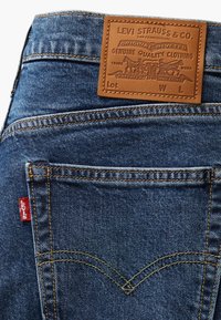 Jean en denim bleu avec un patch en cuir marron embossé d'une marque, une couture orange contrastante et une étiquette de logo rouge sur la poche arrière.