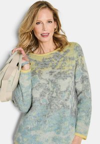 Vrouw met blond haar, gekleed in een meerkleurige grijze, blauwe en gele trui, met een beige tas over haar schouder, en een lichte glimlach.