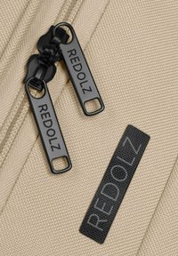 Beige Stoff mit strukturiertem Finish verfügt über zwei schwarze Reißverschlüsse mit der Aufschrift "REDOLZ" und ein schwarzes Textiletikett mit dem Markennamen.