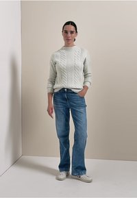 Hellgrauer Kabelstrickpullover kombiniert mit mittelhoch sitzenden blauen Jeans und weißen Sneakers, vor einem neutralen Hintergrund.