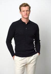 Schwarzer gerippter Langarm-Polo-Pullover mit Kragen und Zwei-Knopf-Blende, kombiniert mit cremefarbenen Hosen. Standardpassform, glatte Textur.