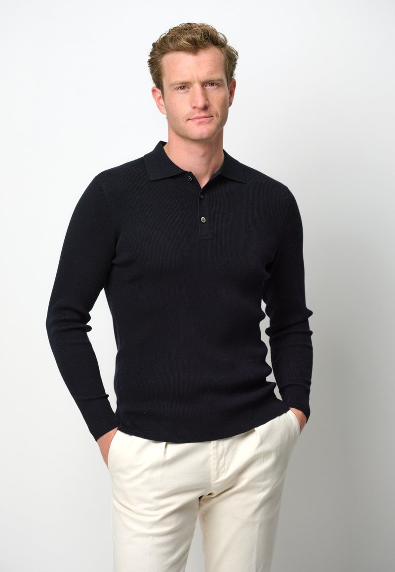 Schwarzer gerippter Langarm-Polo-Pullover mit Kragen und Zwei-Knopf-Blende, kombiniert mit cremefarbenen Hosen. Standardpassform, glatte Textur.