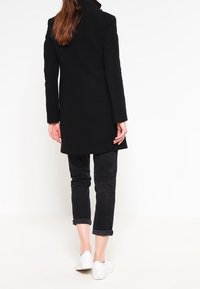 Kvinna stående, iklädd en svart långrock, uppvikta svarta byxor och vita sneakers, vänd bort från en enfärgad vit bakgrund.