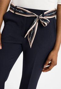 Marineblauwe broek heeft een contrasterende, gestreepte riem van zijde, met een kettingmotief en beige accenten die om de taille zijn gebonden.