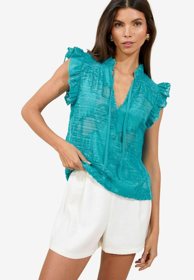 Blusa turchese con maniche a balze e scollatura a V, caratterizzata da un tessuto testurizzato e un motivo floreale, abbinata a pantaloni corti bianchi.