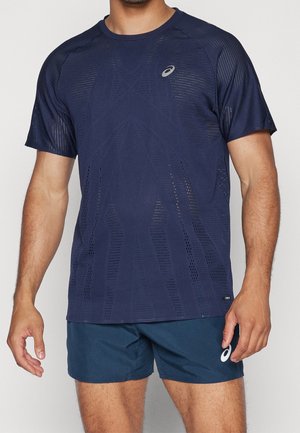 Homme portant un maillot de sport bleu marine perforé et un short assorti bleu marine avec des logos blancs ASICS, debout devant un fond uni.