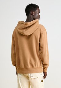 Loose-fitting, hooded sweatshirt in light brown cotton, featuring a soft texture and ribbed cuffs. Simple design with no graphics.