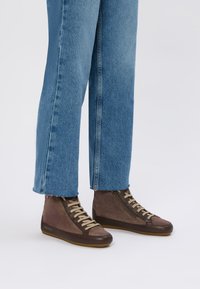 Sneaker alte marroni con una combinazione di suede liscio e strutturato, lacci a contrasto e suola in gomma. Abbinati a jeans blu, leggermente sfilacciati.