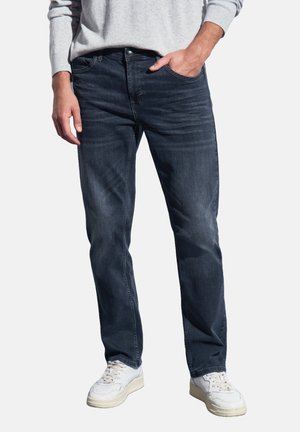 Man in donkere blauwe jeans met witte sneakers en een lichtgrijze trui, staande met één hand in zijn zak op een witte achtergrond.