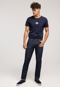 Młody mężczyzna z kręconymi włosami, ubrany w granatowy t-shirt Lee Cooper, ciemne dżinsy i granatowe trampki, stoi z jedną ręką w kieszeni, patrząc w lewo.