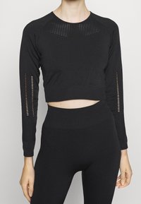 Nainen, jolla on päällään musta saumaton pitkähihaisuuden crop-top ja korkeavyötäröiset legginsit, joissa on geometriset ilmanvaihtorei'itykset hihoissa ja rinnassa.