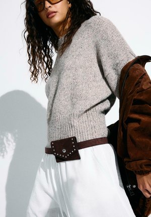 Ženska, ki nosi sivi pleteni pulover, bele hlače, rjavo torbico z zapestjem in drži rjavo jakno čez eno ramo na belem ozadju.