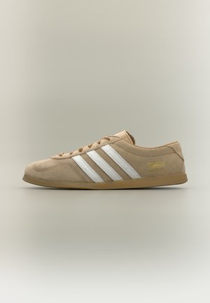 Zapatilla beige Adidas Gazelle con rayas blancas, suela de goma y material de ante mostrada de perfil sobre fondo neutro.